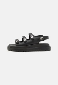 Svart sandal med tre breda remmar med metalldetaljer, en platt sula med mönster och en justerbar bakrem för en säker passform.