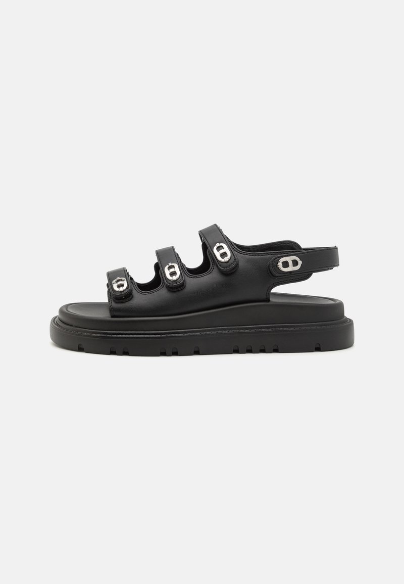 Svart sandal med tre breda remmar med metalldetaljer, en platt sula med mönster och en justerbar bakrem för en säker passform.