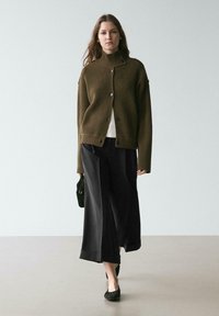 Olivgrüne, gerippte Strickjacke mit hohem Kragen, Knopfleiste und weiten Ärmeln, getragen über einem weißen Top und schwarzen, weit geschnittenen Hosen. Schwarze Ballerinas, kleine Tasche.