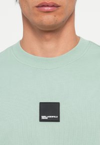 Menta zeleni dukserica s okruglim izrezom izrađena od mekanog materijala, s pravokutnom crnom etiketom s bijelim tekstom "KARL LAGERFELD JEANS" sprijeda.