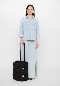 Black wheeled suitcase with silver hardware. Features a smooth texture, adjustable handle, and compact design. Worn by a person in denim outfit.