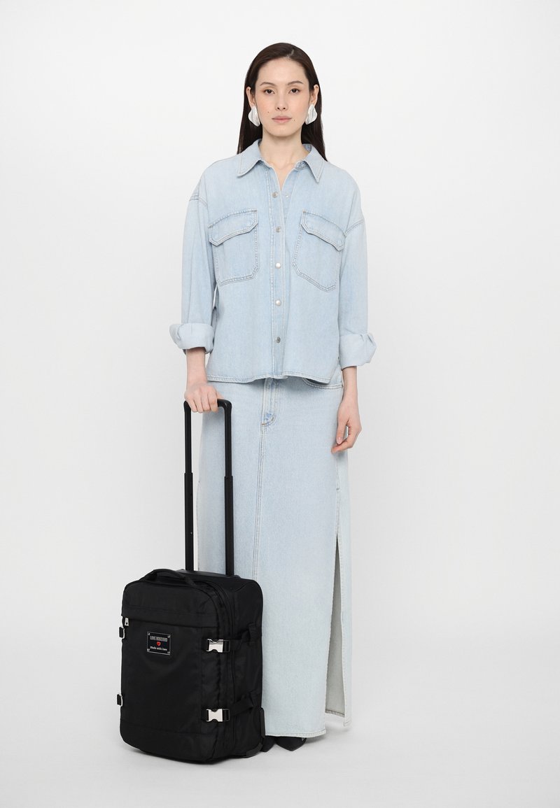 Black wheeled suitcase with silver hardware. Features a smooth texture, adjustable handle, and compact design. Worn by a person in denim outfit.