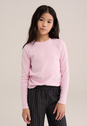Fille aux longs cheveux foncés portant un chemisier rose à manches longues et un pantalon noir à fines rayures, debout devant un fond neutre.