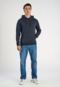 Felpa navy con tasca frontale, cappuccio con cordino e polsini a costine, abbinata a jeans blu e scarpe da ginnastica bianche, su uno sfondo neutro.