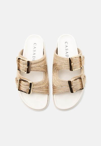 Paire de sandales blanches à brides beiges texturées et grandes boucles dorées, nom de la marque "CASADEI" visible sur les semelles.