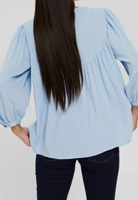 Blouse bleu clair à manches bouffantes, en tissu texturé et à coupe décontractée. Le dos présente une couture en yoke pour un détail de conception supplémentaire.