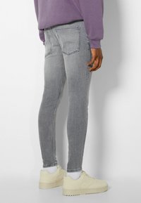 Hombre vistiendo jeans skinny gris claro, calcetines blancos, zapatillas crema y una sudadera morada, de pie contra un fondo blanco.