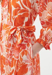 Abito floreale con stampa arancione e rosa, vita accentuata con nastro in tessuto, maniche lunghe con polsini arricciati e dettagli con bottoni.