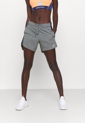 Sportowiec noszący szare spodenki Nike ze sznurkiem, pomarańczowy i granatowy sportowy stanik Nike Pro oraz białe buty sportowe, stojący na białym tle.