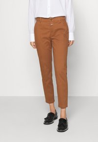 Brązowe bawełniane spodnie o wąskim kroju, zapinane na guziki, z przednimi kieszeniami, zestawione z czarnymi wsuwanymi butami i białą koszulą na guziki.