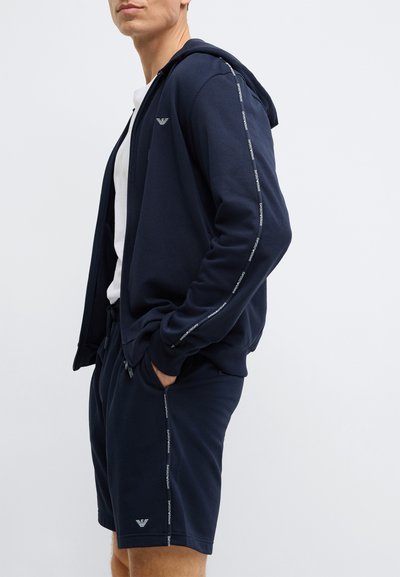 Tengerészkék cipzáras kapucnis pulóver logóval a mellkasán és függőleges szöveges díszítéssel az ujjain, hozzá illő rövidnadrággal, amelyen oldalsó logók találhatók.