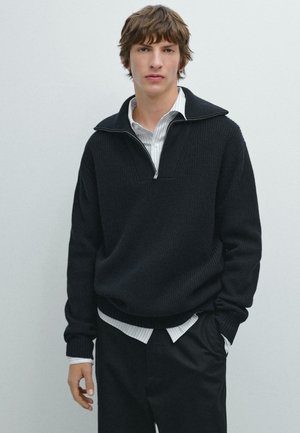 Pullover nero lavorato a maglia con colletto con zip, indossato sopra una camicia bianca a righe. Il pullover ha una vestibilità ampia e polsini a coste.