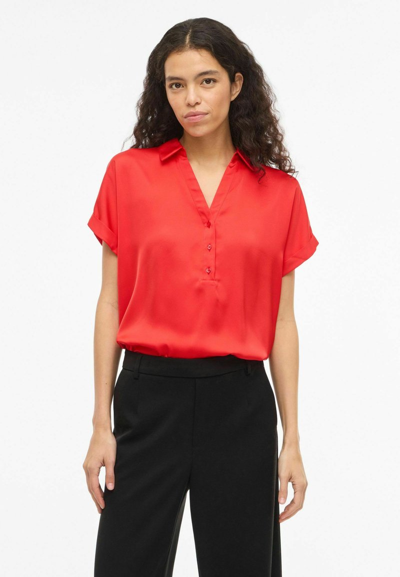 Frau mit lockigem, dunklem Haar, die eine leuchtend rote Bluse mit kurzen Ärmeln und hoch taillierte schwarze Hose trägt, vor einem weißen Hintergrund stehend.