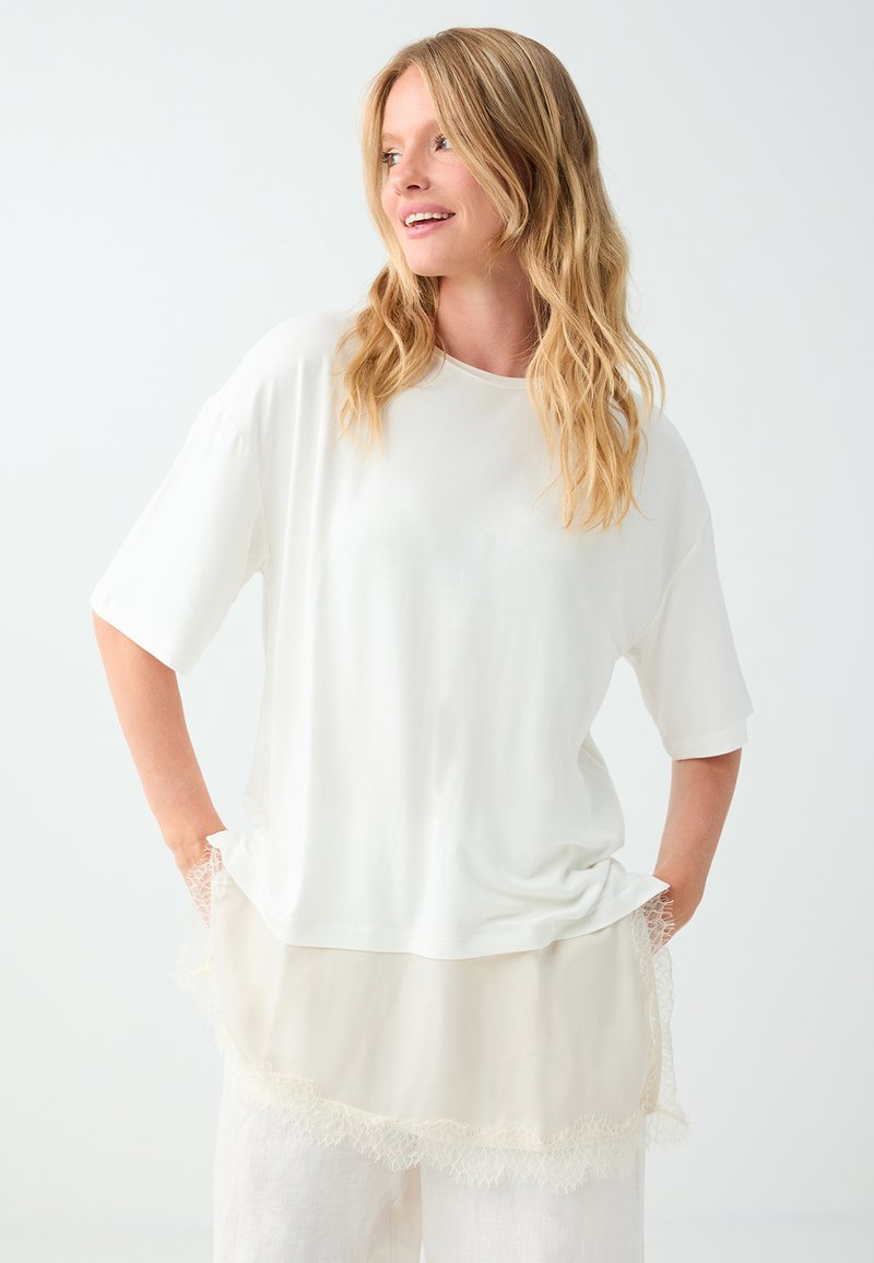 Femme blonde portant un haut blanc ample à manches courtes et une jupe crème à volants bordée de dentelle, souriante et regardant vers sa gauche, les mains dans les poches.
