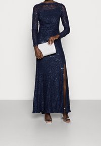Navy lace evening gown with sequins, long sleeves, and thigh-high slit. Paired with a silver clutch and clear heeled sandals.