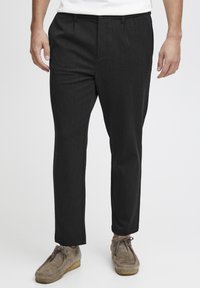 Pantalons noirs ajustés avec une texture lisse, devant plat et poches latérales. Associés à des chaussures en style mocassins grises.