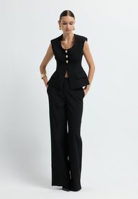 Gilet nero su misura con scollatura a forma di U, tre bottoni dorati e tasche laterali, abbinato a pantaloni neri a vita alta. Texture liscia.