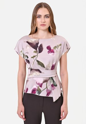 Donna con capelli lunghi che indossa una camicetta floreale viola chiaro con maniche corte e pantaloni neri su sfondo bianco.