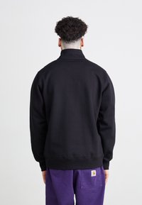 Sort sweatshirt med høj krave, ribstrikkede manchetter og kant. Den har en glat tekstur og et enkelt design, båret med lilla bukser.