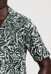 Camicia a maniche corte con colletto in tessuto strutturato, caratterizzata da un motivo di foglie verdi e bianche con linee spesse e forme organiche.