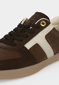 Καφέ και μπεζ αθλητικό παπούτσι Tommy Hilfiger με μαύρα κορδόνια και χρυσά ρουμπάκια, με λευκή λωρίδα στο πλάι και λαστιχένια σόλα.