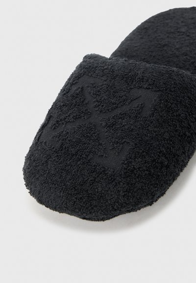 Chausson en tissu éponge noir avec motif géométrique texturé sur l\'orteil, posé sur un fond clair uni.