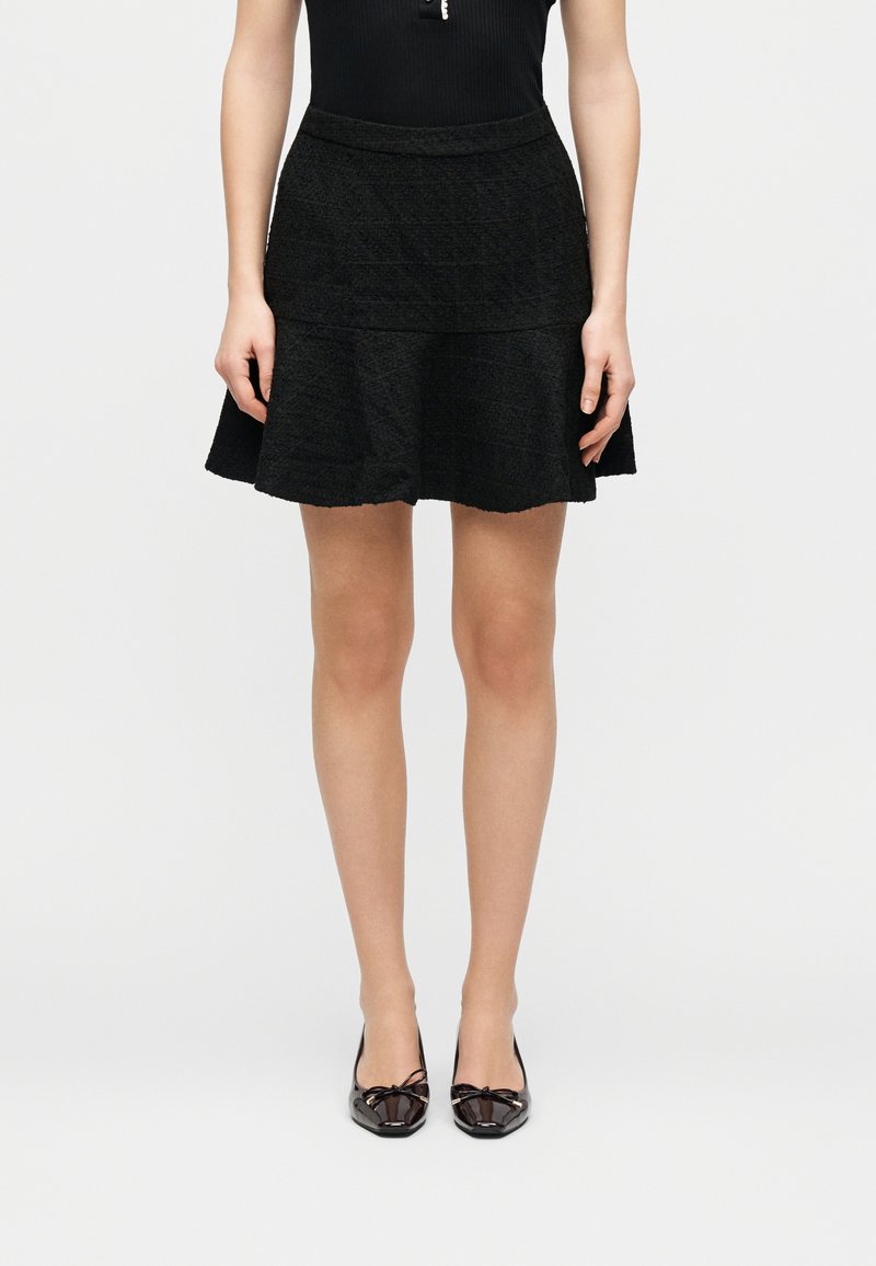 Γυναίκα που φοράει κοντή μαύρη υφασμάτινη φούστα, μαύρο αμάνικο τοπ και μαύρες μπαλαρίνες από δερματίνη με φιόγκους, που στέκεται σε λευκό φόντο.