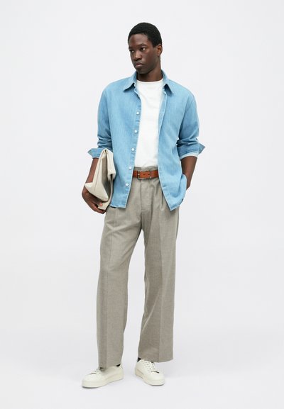 Jovem com calças bege, ténis brancos, camisa branca e camisa de ganga azul, segurando uma bolsa bege dobrada contra um fundo simples.