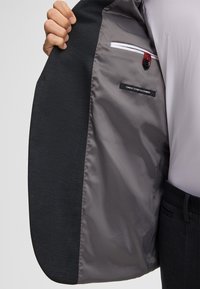 Uomo in abito scuro solleva la giacca per rivelare l'interno grigio con tasca abbottonata e etichetta "Finest Stretch Fabric".