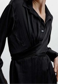 Robe longue noire et légère à manches longues avec col, fermeture à boutons et ceinture nouée à la taille, texture lisse.