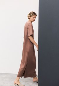 Brun klänning i ankellängd med korta ärmar och en sidospalt. Mjukt, slätt tyg med avslappnad passform. Modellen står mot en grå vägg.