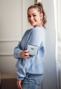 Light blue ribbed sweater, gray jeans, holding a light blue textured wallet with a purple trim and embossed logo. Natural light background.