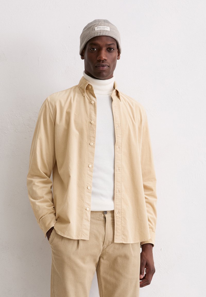 Chemise en velours côtelé beige avec des boutons, portée par-dessus un col roulé blanc. Bonnet en tricot gris avec une étiquette. Le mannequin se tient devant un fond uni.