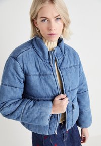 Blue padded jacket with a high collar, featuring a zip front and subtle stitching. Worn over a light sweater, paired with a denim skirt.