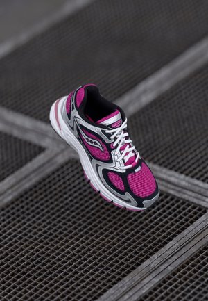 Zapatilla de correr Saucony en colores rosa, negro y blanco con cordones blancos, descansando sobre una superficie metálica con rejilla.