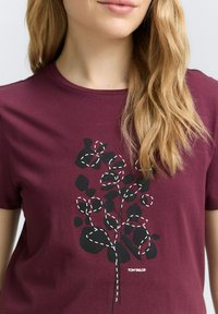 T-shirt court à manches courtes de couleur bordeaux en coton, avec un design botanique noir et des accents de couture blanche, et le logo "TOM TAILOR" en bas.