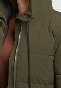 Olivengrøn pufferjakke med høj krave, der har en lynlåsfront og sort snor. Stoffet ser glat ud med quiltede detaljer.