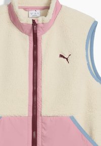 Fleece-vesti, jossa on creamy-väriinen runko, vaaleanpunainen etuvetoketju ja sininen reunus. Vasemmassa rinnassa on pieni ruskea logo. Pehmeä rakenne, hihaton malli.