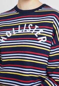 Marinrandig sweatshirt med röda, gula och ljusblå linjer. Har vit text "HOLLISTER" över bröstet och en rundad halsringning.