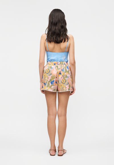 Γυναίκα με σκούρα μαλλιά μέχρι τους ώμους, που φορά μπλε τοπ με λεπτή τιράντα και σορτς με λουλουδάτο μοτίβο, στέκεται γυρισμένη με την πλάτη.