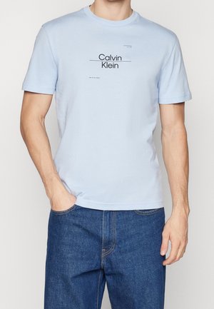 Muž v světle modrém tričku Calvin Klein a tmavě modrých džínách s jednou rukou v kapse stojí před jednoduchým pozadím.