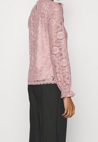 Top lungo rosa in pizzo con maniche a sbuffo e orlo ondulato, decorato con motivi floreali e polsini arricciati. Presenta un'apertura a chiave sul retro. Indossato con pantaloni scuri.