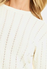 Suéter blanco de punto con un patrón texturizado que presenta rayas verticales y formas de rombo, cuello redondo y mangas holgadas.