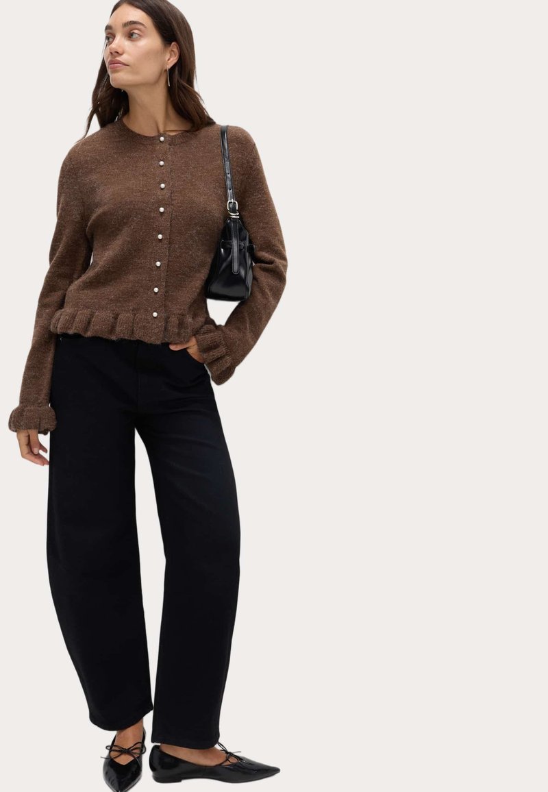 Kobieta nosi brązowy kardigan zapinany na guziki z falbaniastymi brzegami, czarne spodnie w szerokim kroju, czarne baleriny oraz trzyma małą czarną torebkę na ramieniu.