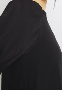 Abito nero con dettagli di maniche a palloncino mostrato sul torso superiore accanto a uno sfondo chiaro, orecchini con gemme multicolori visibili.
