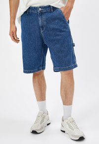 Pantalones cortos de mezclilla en azul oscuro con un corte recto, bolsillos laterales y dobladillo enrollado. Llevados con zapatillas blancas y calcetines.