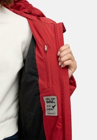 Red insulated jacket with a zip pocket inside. Features a fabric label stating "We are one. Peace Clothing." Textured black lining.