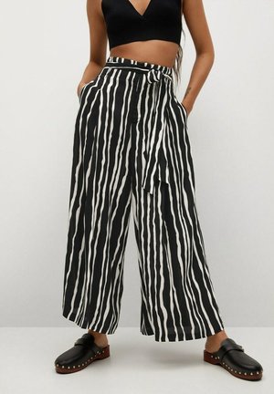 Femme portant un pantalon à larges jambes rayé verticalement noir et blanc avec une ceinture nouée, un crop top noir et des sabots noirs à enfiler.