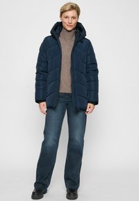 Frau trägt eine marineblaue gepolsterte Kapuzenjacke, einen braunen Rollkragenpullover, dunkelblaue Jeans und schwarze Schuhe und steht vor einem hellen Hintergrund.