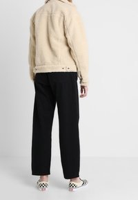 Veste en shearling beige avec col, coutures visibles et boutons-pression sur les côtés, associée à un pantalon large noir et des mocassins à carreaux.